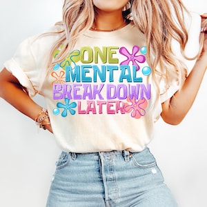 May include: A white t-shirt with a graphic design featuring colorful flowers and the text "One Mental Break Down Later".