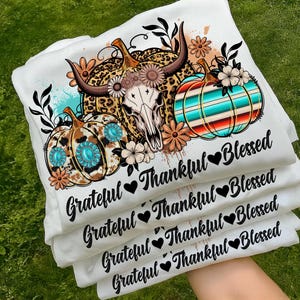Puede incluir: Camisetas blancas apiladas, con un dise&ntilde;o tem&aacute;tico de oto&ntilde;o. El dise&ntilde;o incluye calabazas, una calavera de vaca y elementos florales. El texto en las camisetas dice "Grateful, Thankful, Blessed" con s&iacute;mbolos de coraz&oacute;n. Las calabazas tienen patrones turquesa y marr&oacute;n.