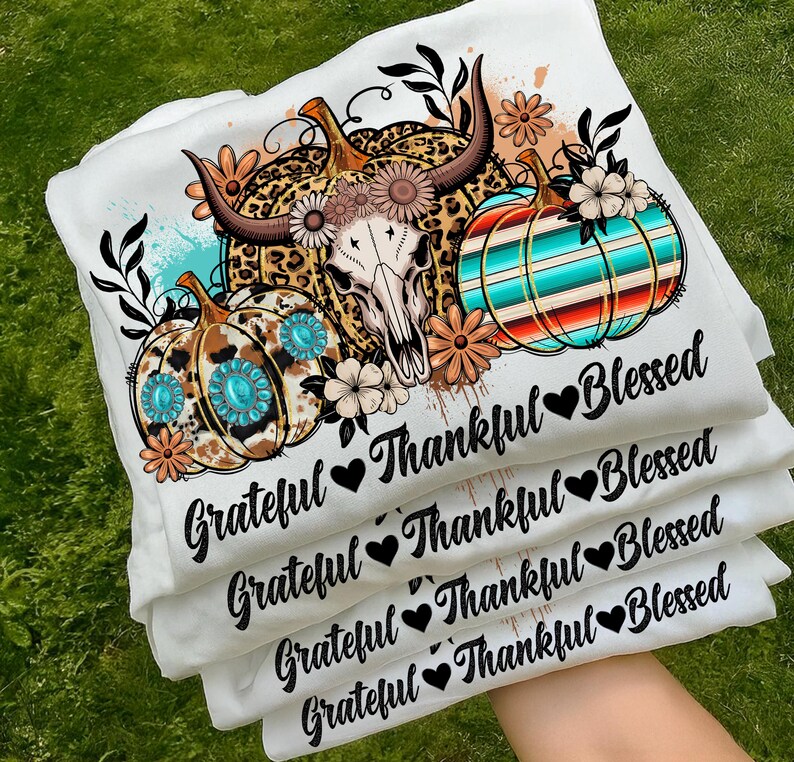 Puede incluir: Camisetas blancas apiladas, con un dise&ntilde;o tem&aacute;tico de oto&ntilde;o. El dise&ntilde;o incluye calabazas, una calavera de vaca y elementos florales. El texto en las camisetas dice "Grateful, Thankful, Blessed" con s&iacute;mbolos de coraz&oacute;n. Las calabazas tienen patrones turquesa y marr&oacute;n.