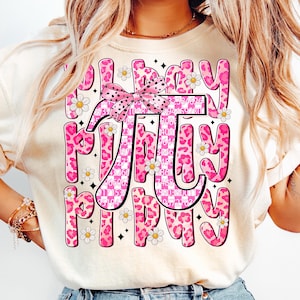 Puede incluir: Una camiseta blanca con un diseño rosa y blanco que dice "Pi Day" con un lazo rosa y flores blancas. La "Pi" está hecha de un estampado de leopardo rosa con números en su interior.