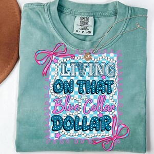 Pu&ograve; includere: T-shirt Comfort Colors verde acqua chiaro con la scritta "LIVING ON THAT Blue Collar DOLLAR" in un design decorativo. Il design include fiocchi rosa, stelle e uno sfondo a scacchi blu e bianco. Una collana d'argento &egrave; drappeggiata sulla maglietta.