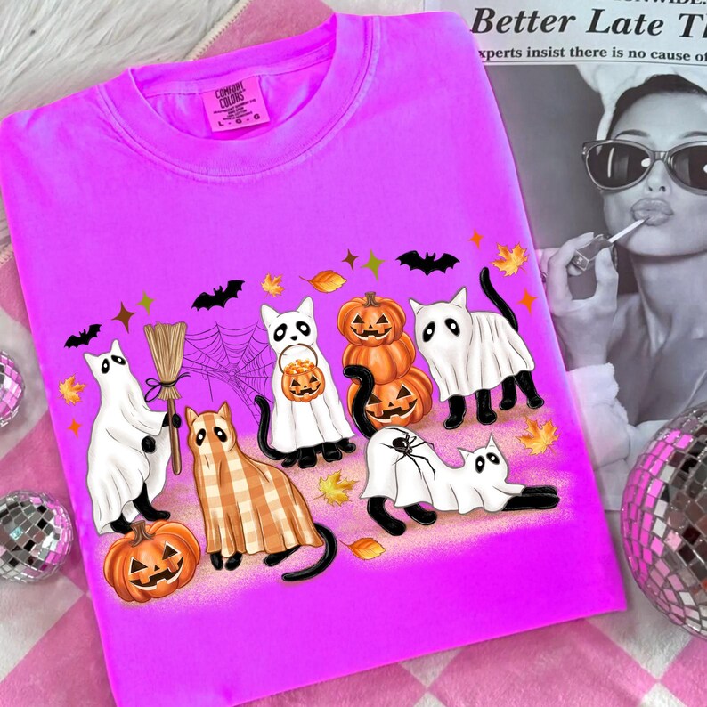 May include: A vibrant pink Comfort Colors t-shirt with a Halloween graphic. The design features cartoon cats in ghost costumes, pumpkins, bats, and a spider web. The shirt has a crew neck and a Comfort Colors label.