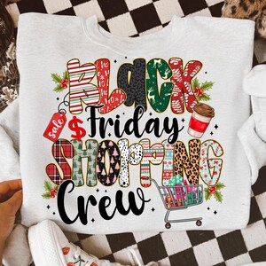 May include: A light gray sweatshirt with a colorful graphic that reads "Black Friday Shopping Crew." The text incorporates various patterns, including stripes, leopard print, and holiday designs. A shopping cart filled with gifts is also featured.