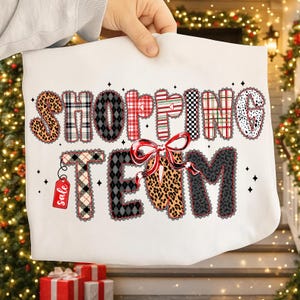 Peut inclure: Un sac fourre-tout blanc avec l'inscription "SHOPPING TEAM" dans un design patchwork. Les lettres pr&eacute;sentent des imprim&eacute;s l&eacute;opard, des carreaux et un n&oelig;ud rouge. Une petite &eacute;tiquette rouge avec le mot "sale" est suspendue &agrave; la lettre "T".
