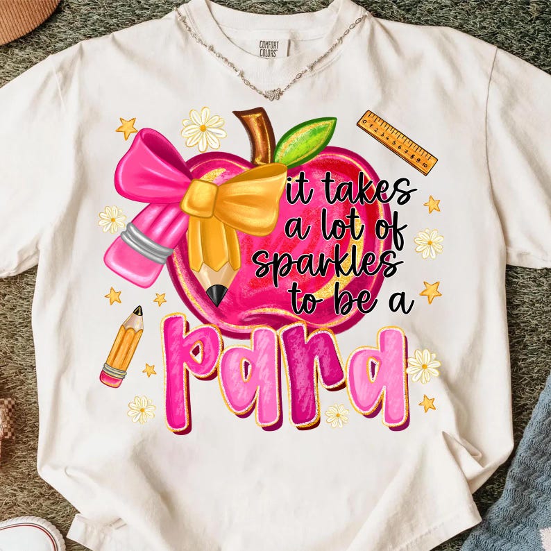 May include: White t-shirt featuring a pink apple design with a pencil, bow, and the text "it takes a lot of sparkles to be a para". Additional details include a ruler, stars, and flowers.