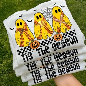 Puede incluir: Varias camisetas blancas apiladas, con un dise&ntilde;o tem&aacute;tico de Halloween. El dise&ntilde;o incluye figuras de fantasmas amarillos con pelotas de b&eacute;isbol, bates y guantes, junto con calabazas y murci&eacute;lagos. El texto "Tis The season" est&aacute; impreso en negro.