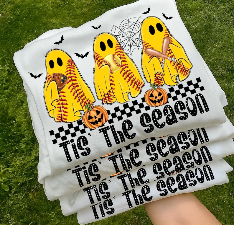 Puede incluir: Varias camisetas blancas apiladas, con un dise&ntilde;o tem&aacute;tico de Halloween. El dise&ntilde;o incluye figuras de fantasmas amarillos con pelotas de b&eacute;isbol, bates y guantes, junto con calabazas y murci&eacute;lagos. El texto "Tis The season" est&aacute; impreso en negro.