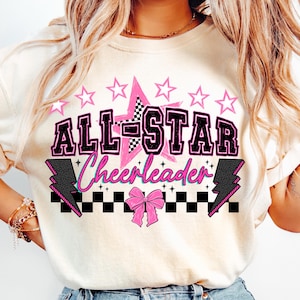 May include: Cream-colored sweatshirt with a graphic that says "ALL-STAR Cheerleader" in black and pink. The design includes a pink star, pink stars, a checkered pattern, and lightning bolts. A pink bow is at the bottom of the design.