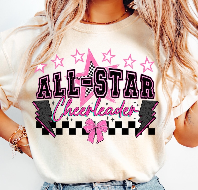 May include: Cream-colored sweatshirt with a graphic that says "ALL-STAR Cheerleader" in black and pink. The design includes a pink star, pink stars, a checkered pattern, and lightning bolts. A pink bow is at the bottom of the design.