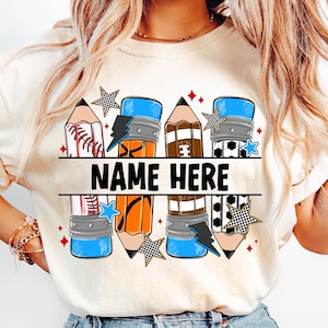 Pode incluir: Camiseta creme com um design de l&aacute;pis que incorpora elementos esportivos. O design inclui gr&aacute;ficos de beisebol, basquete e futebol, juntamente com o texto "NAME HERE". Os detalhes incluem estrelas e raios.