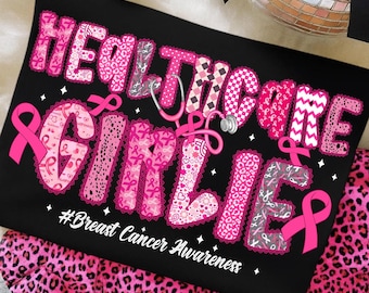 May include: Black t-shirt with the words "Healthcare Girlies" in pink, decorated with pink ribbons and a stethoscope. The phrase "#Breast Cancer Awareness" is also printed on the shirt. The shirt is placed on a pink leopard print fabric.