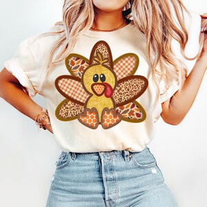 May include: A white t-shirt featuring a cute cartoon turkey wearing a patchwork costume. The turkey is brown, yellow, and orange with a red beak and wattle. The patchwork costume is made of different fabrics, including plaid, leopard print, and floral.
