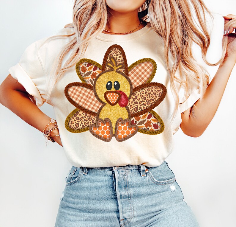May include: A white t-shirt featuring a cute cartoon turkey wearing a patchwork costume. The turkey is brown, yellow, and orange with a red beak and wattle. The patchwork costume is made of different fabrics, including plaid, leopard print, and floral.