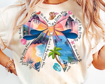 May include: A white t-shirt with a colorful bow design. The bow is made up of beach scenes and the text "The Beach Is My Happy Place" is repeated around the bow.