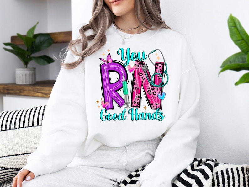 May include: White sweatshirt with a pink, purple, and blue graphic that says "You RN Good Hands". The graphic includes a stethoscope, a butterfly, and a syringe.