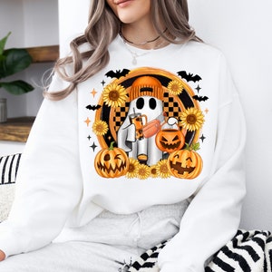 Peut inclure: Sweat-shirt blanc avec un motif sur le th&egrave;me d'Halloween. Le motif comprend un fant&ocirc;me portant un bonnet orange, tenant une boisson et une lanterne citrouille. Des tournesols, des chauves-souris et des &eacute;toiles entourent le fant&ocirc;me et les citrouilles.