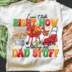 May include: White t-shirt with a colorful graphic featuring a barbecue scene. Text reads "I can't talk RIGHT NOW I'm doing hot DAD STUFF." The design includes a grill, beer, lawnmower, and food.