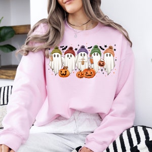 Op de afbeelding: Roze sweatshirt met een afbeelding van vijf geesten die hoeden dragen en pompoenen en koffiebekers vasthouden. De geesten dragen kleurrijke hoeden en glimlachen. De afbeelding is omringd door vleermuizen en sterren.