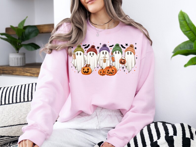 Op de afbeelding: Roze sweatshirt met een afbeelding van vijf geesten die hoeden dragen en pompoenen en koffiebekers vasthouden. De geesten dragen kleurrijke hoeden en glimlachen. De afbeelding is omringd door vleermuizen en sterren.