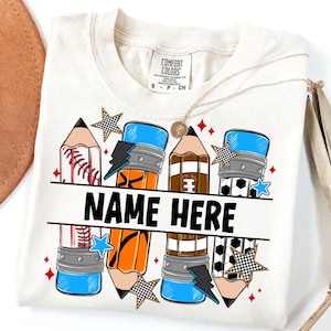 Pode incluir: Camiseta creme com um design de l&aacute;pis com temas de beisebol, futebol americano e futebol. A camisa inclui o texto "NAME HERE" em letras pretas em negrito, com estrelas e raios. Um colar de ouro est&aacute; drapeado sobre a camisa.