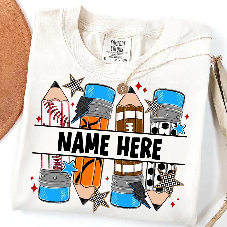 Pode incluir: Camiseta creme com um design de l&aacute;pis com temas de beisebol, futebol americano e futebol. A camisa inclui o texto "NAME HERE" em letras pretas em negrito, com estrelas e raios. Um colar de ouro est&aacute; drapeado sobre a camisa.