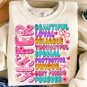 Op de afbeelding: Een wit sweatshirt met het woord "Zus" gespeld in kleurrijke letters met bloemen en andere decoratieve elementen. De letters worden omringd door de woorden "Mooi, Loyal, Betrouwbaar, Gedachtzaam, Speciaal, Beschermend, Uniek, Beste vriend, Voor altijd".