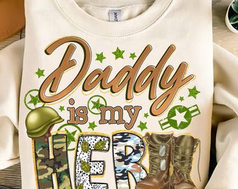 Könnte beinhalten: Ein weißer Pullover mit dem Text "Daddy is my Hero" in goldenen Buchstaben. Der Text ist von grünem Tarnmuster und Sternen umgeben. Am unteren Rand des Pullovers befinden sich zwei braune Stiefel mit grünen Schnürsenkeln und Hundemarken.