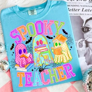 May include: A light blue t-shirt with the words "Spooky Teacher" in colorful block letters. The shirt features three cartoon ghosts reading books, with bats and spiderwebs. The ghosts are in pink, blue, and yellow.