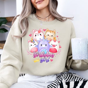 Puede incluir: Una sudadera beige con un gr&aacute;fico de cinco animales de dibujos animados, incluyendo un unicornio, un gato, una llama y dos criaturas m&aacute;s. El texto "birthday girl" est&aacute; impreso debajo de los animales.