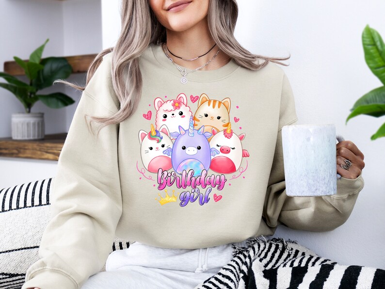 Puede incluir: Una sudadera beige con un gr&aacute;fico de cinco animales de dibujos animados, incluyendo un unicornio, un gato, una llama y dos criaturas m&aacute;s. El texto "birthday girl" est&aacute; impreso debajo de los animales.