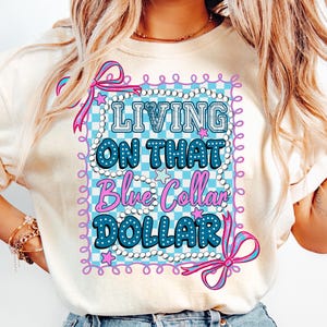 Pu&ograve; includere: T-shirt color crema con una grafica che recita "LIVING ON THAT Blue Collar DOLLAR" in blu e rosa. Il design presenta uno sfondo a scacchi, stelle e bordi decorativi.