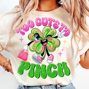 May include: A white t-shirt with a green shamrock design. The shamrock has a pink face with a wink and a smile. The text "Too Cute To Pinch" is written in pink and green letters.