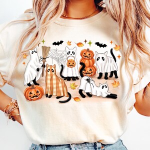 May include: A cream-coloured sweatshirt with a Halloween design. The graphic features cartoon ghosts, cats, pumpkins, bats, and autumn leaves. The design uses orange, black, white, and brown.