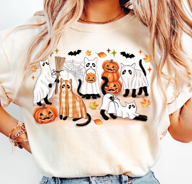 May include: A cream-coloured sweatshirt with a Halloween design. The graphic features cartoon ghosts, cats, pumpkins, bats, and autumn leaves. The design uses orange, black, white, and brown.