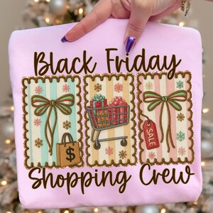 May include: A light pink sweatshirt with the words "Black Friday Shopping Crew" in brown script. The design includes three framed illustrations with bows, a shopping cart, a gift bag, and a sale tag.