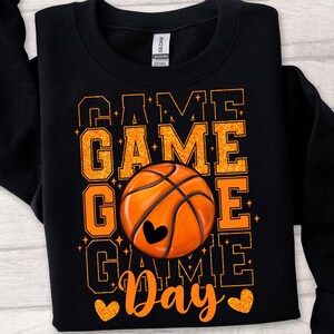 Peut inclure: Sweat-shirt noir avec un texte en paillettes orange qui dit "Game Game Go Game Day" avec un graphique de ballon de basket au centre.