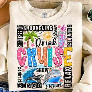 May include: A beige sweatshirt with a colorful graphic design featuring the word "CRUISE" surrounded by words like "Drink", "Bingo", "Swim", "Relax", "Show", "Ocean", "Snorkeling", "Islands", and "Sightseeing".