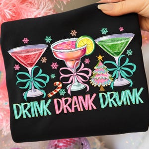 May include: Black sweatshirt with a festive design featuring three cocktail glasses, a candy cane, and a Christmas tree. The text "DRINK DRANK DRUNK" is displayed in colorful letters. The glasses are decorated with bows and snowflakes.