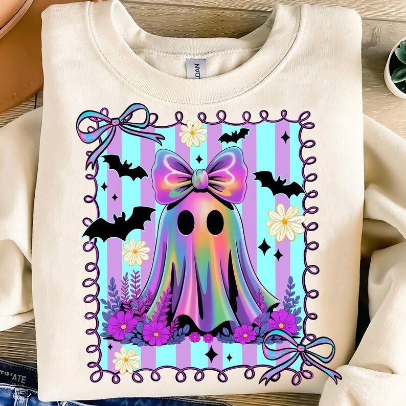 May include: Cream-colored sweatshirt featuring a colorful ghost design. The ghost has a large bow and is surrounded by bats, flowers, and a decorative border. The design is set against a striped background of blue and purple.
