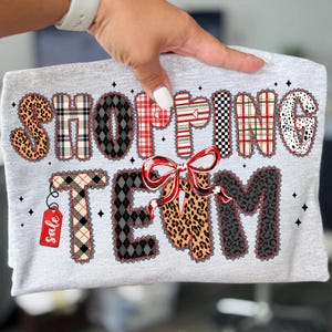 Peut inclure: Un t-shirt gris clair avec l'inscription "SHOPPING TEAM" dans diff&eacute;rents motifs, notamment &agrave; carreaux, l&eacute;opard et pois. Un n&oelig;ud rouge et une &eacute;tiquette "soldes" sont &eacute;galement pr&eacute;sents dans le design.