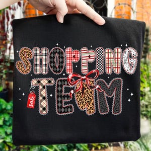 Peut inclure: Sweat-shirt noir avec l'inscription "SHOPPING TEAM" dans un motif patchwork. Les lettres sont d&eacute;cor&eacute;es d'imprim&eacute;s l&eacute;opard, &eacute;cossais et &agrave; pois. Un n&oelig;ud rouge et une &eacute;tiquette "soldes" ajoutent des d&eacute;tails festifs.
