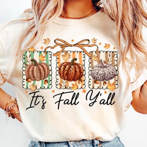 May include: Cream-colored sweatshirt featuring a fall-themed graphic. The design includes three framed pumpkins with different patterns, surrounded by leaves and acorns. The text "It's Fall Y'all" is written in a script font below the pumpkins.