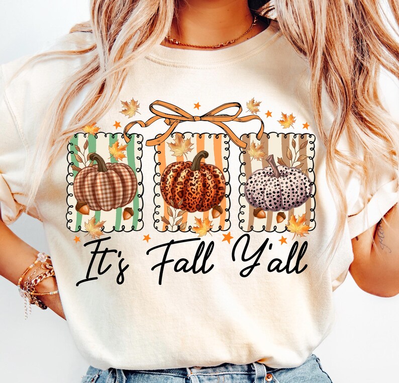 May include: Cream-colored sweatshirt featuring a fall-themed graphic. The design includes three framed pumpkins with different patterns, surrounded by leaves and acorns. The text "It's Fall Y'all" is written in a script font below the pumpkins.