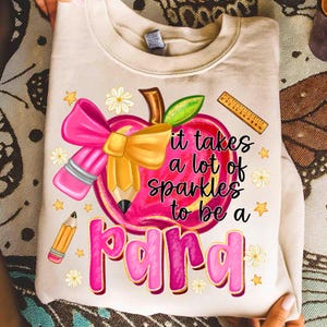 May include: Cream-colored sweatshirt with a graphic featuring a pink apple with a pencil, bow, and the text "it takes a lot of sparkles to be a para." Additional elements include a ruler, stars, and flowers.