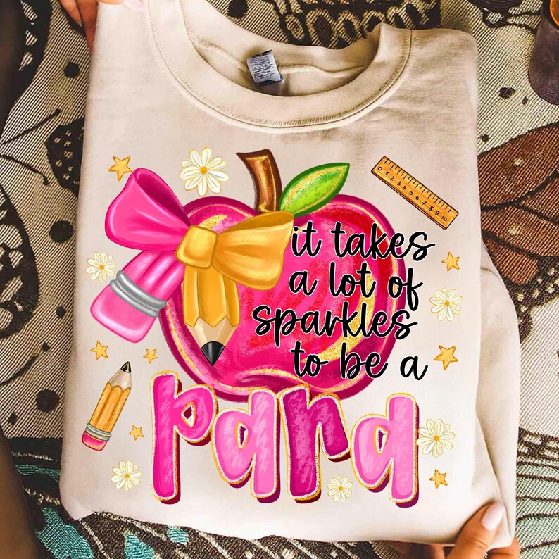 May include: Cream-colored sweatshirt with a graphic featuring a pink apple with a pencil, bow, and the text "it takes a lot of sparkles to be a para." Additional elements include a ruler, stars, and flowers.