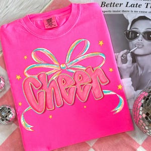 May include: A bright pink t-shirt with the word "Cheer" in a bubble font, outlined in gold, and a colorful bow design. The shirt also features small yellow star accents. The shirt is made of Comfort Colors material.
