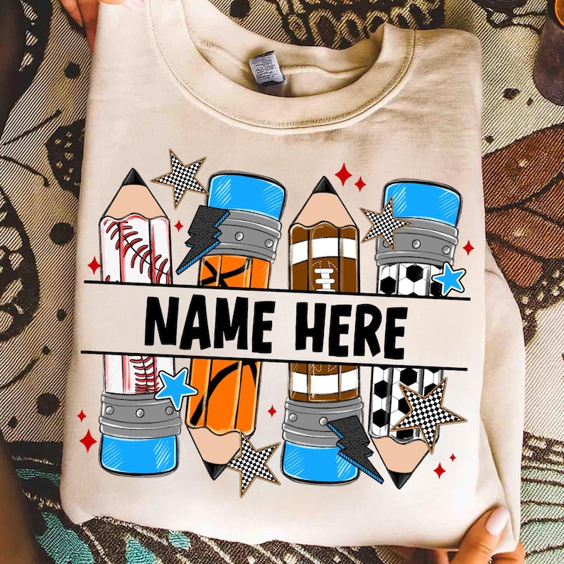 Pode incluir: Camisola bege com um design de l&aacute;pis que incorpora elementos de basebol, basquetebol, futebol americano e bola de futebol. O design inclui o texto "NAME HERE" em negrito preto, juntamente com estrelas e raios.