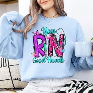 May include: A light blue sweatshirt with the text "You RN Good Hands" in pink, purple, and black lettering. The letters are decorated with a stethoscope, a butterfly, a syringe, and leopard print.