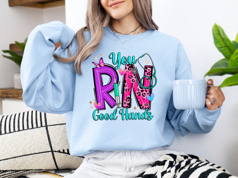 May include: A light blue sweatshirt with the text "You RN Good Hands" in pink, purple, and black lettering. The letters are decorated with a stethoscope, a butterfly, a syringe, and leopard print.