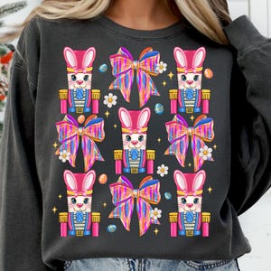 Puede incluir: Una sudadera negra con un patrón repetido de figuras de cascanueces de conejo rosa y lazos coloridos. Los conejos llevan uniformes azules y blancos y tienen orejas y narices rosas.